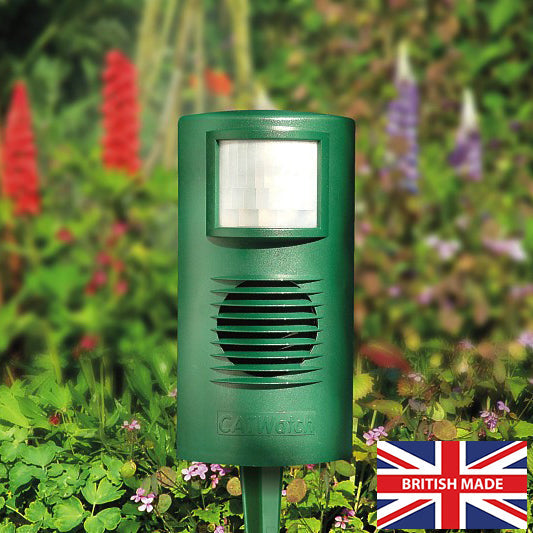 An ultrasonic cat deterrent device with a green housing, emitting a beam of light, placed in a garden setting with flowers in the background.