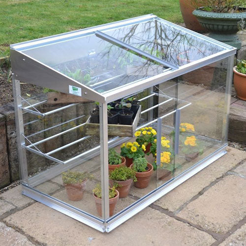 A small Access Half Growhouse with glass on all sides and sliding doors, placed on a garden paving with potted plants inside.