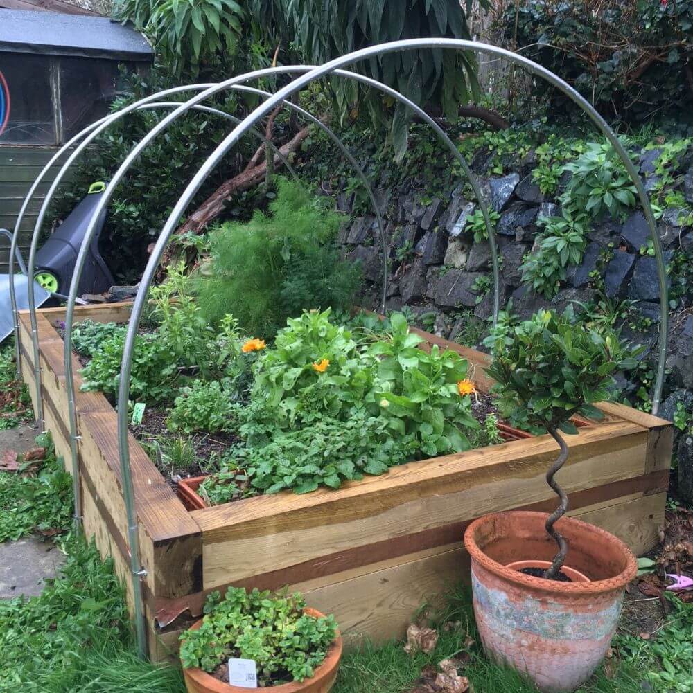 Wooden raised garden bed with metal arch supported by a stone wall and greenery.