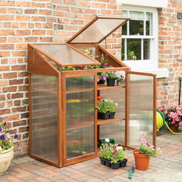 A Rowlinson Hardwood Mini Greenhouse with twin-wall polycarbonate glazing and metal lid stays, situated on a brick pathway next to a wall.