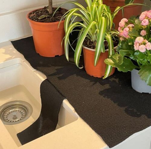 A black capillary matting used for watering plants, placed on a sink with potted plants in the background.