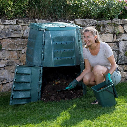 A woman crouching next to a green Thermo Star compost bin, which is full of garden waste, and a black bin with a lid, placed on a grassy area.