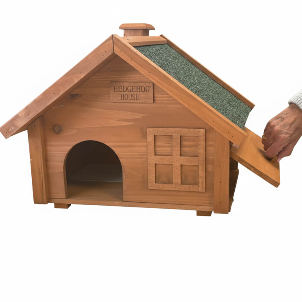 A small wooden hedgehog house with a slanted roof and a hand lifting the outer cover flap, shown on a plain white background.