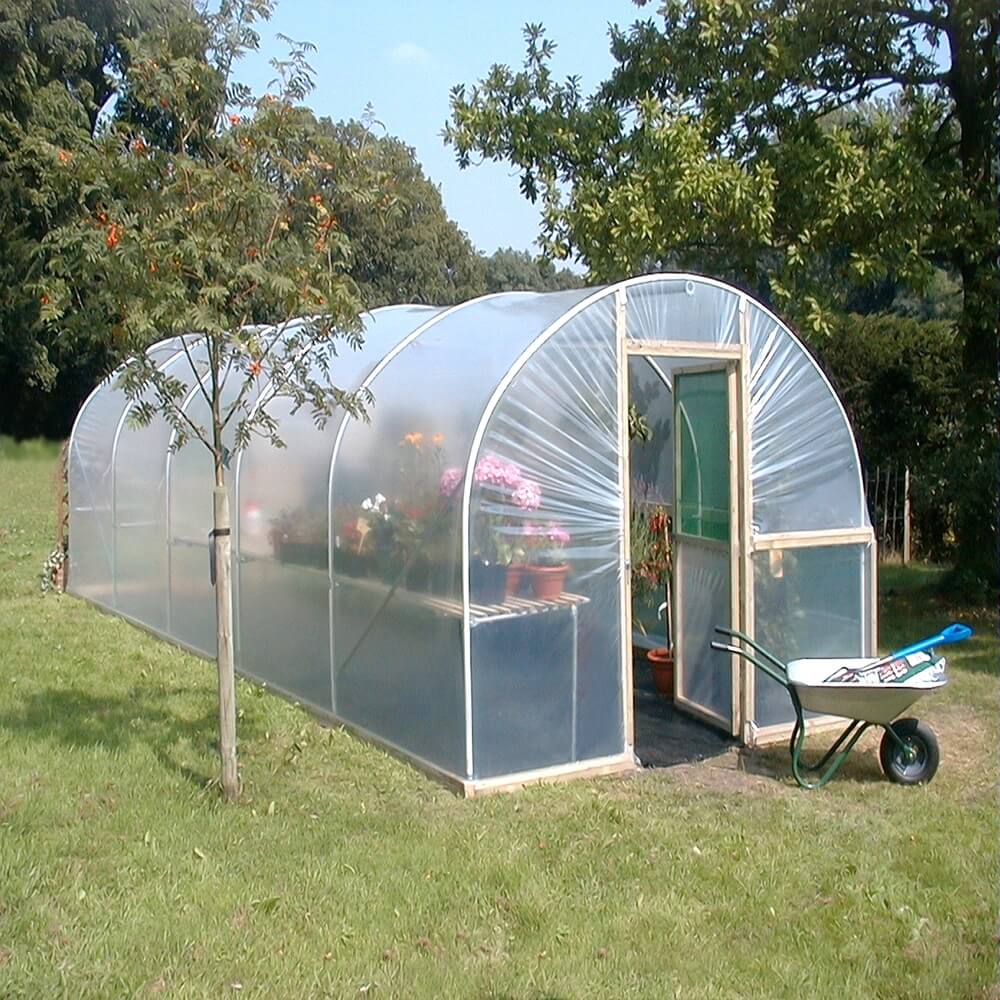 Polycarbonate greenhouse with plants and a wheelbarrow on a grassy area with trees in the background