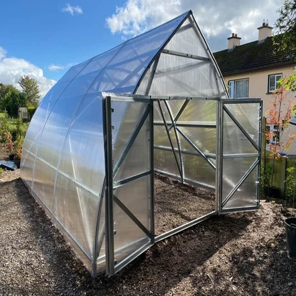 Greenhouse with transparent roof and clear walls on a gravel surface with a house in the background.