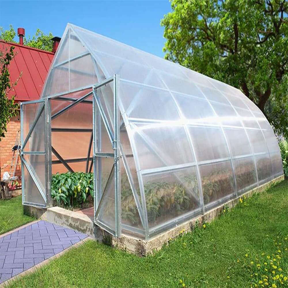 Greenhouse with transparent polycarbonate panels on a grassy area with trees and a building in the background.