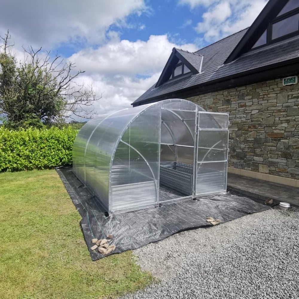 Greenhouse with a metallic frame and clear polycarbonate covering, situated outdoors with a fence in the background.