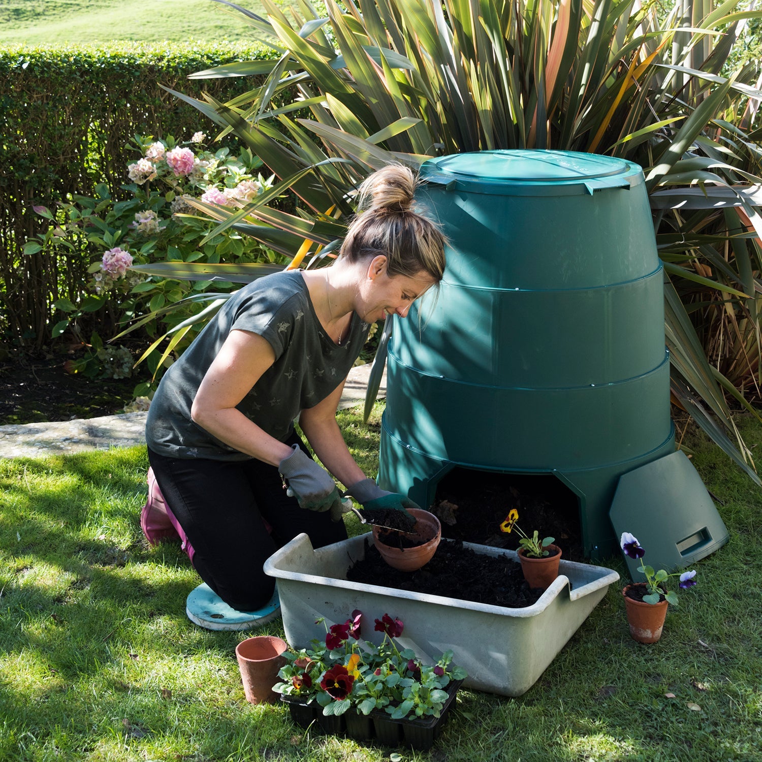 A person crouching next to a green Johanna hot composter, which is filled with various potted plants. 