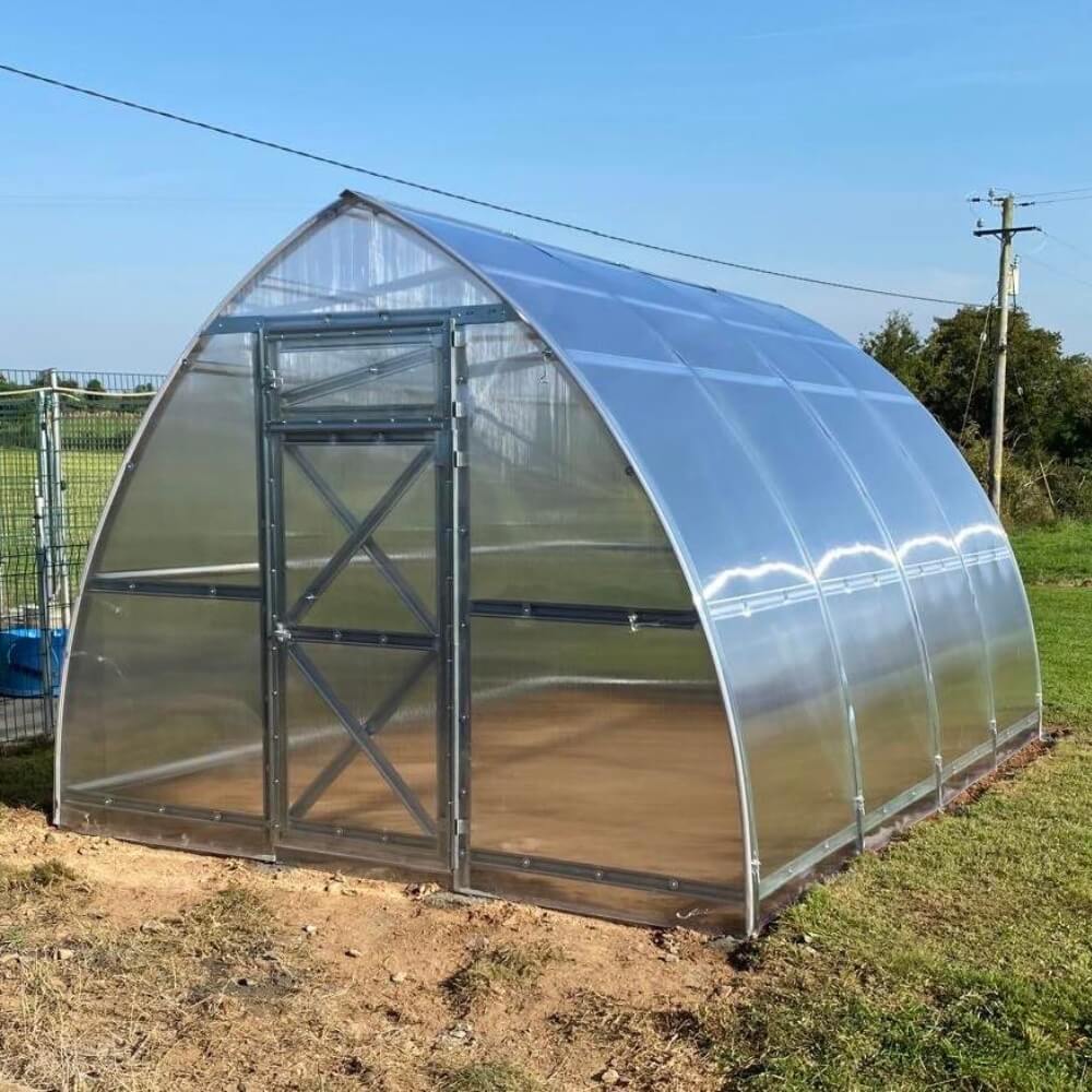 Polycarbonate greenhouse with a metal door on a grassy field under a clear blue sky.