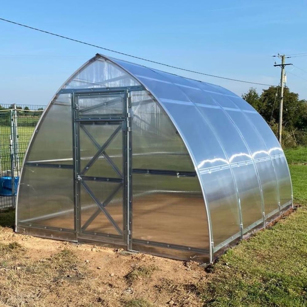 Polycarbonate greenhouse with metal door on a grassy field under a clear blue sky.