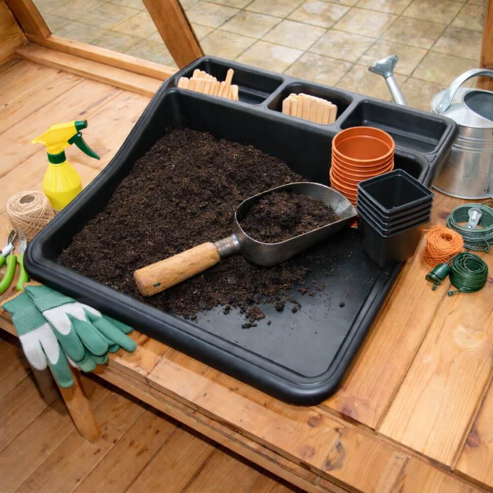 Gardening tools and supplies on a wooden table including a black tray with soil, a scoop, gloves, and pots.