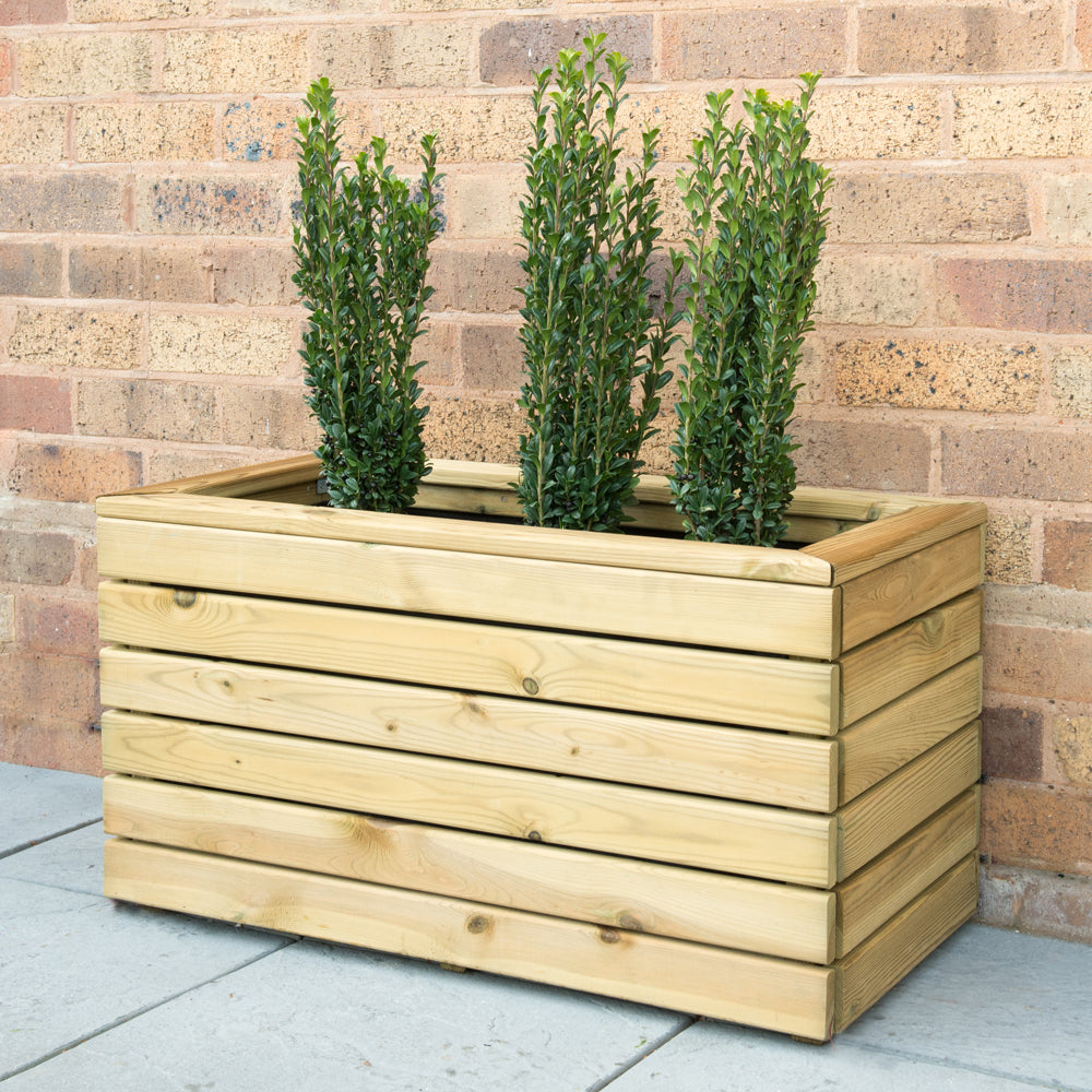 A rectangular wooden planter with a porous liner, containing several shrubs, placed against a brick wall on a concrete path.